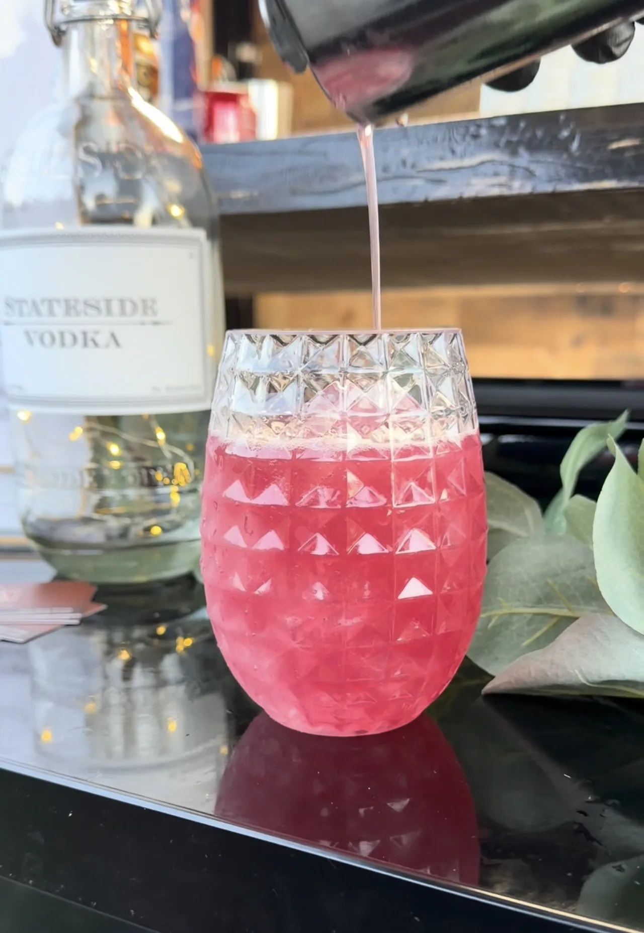 A pink cocktail being poured from a shaker into a textured glass at the bar