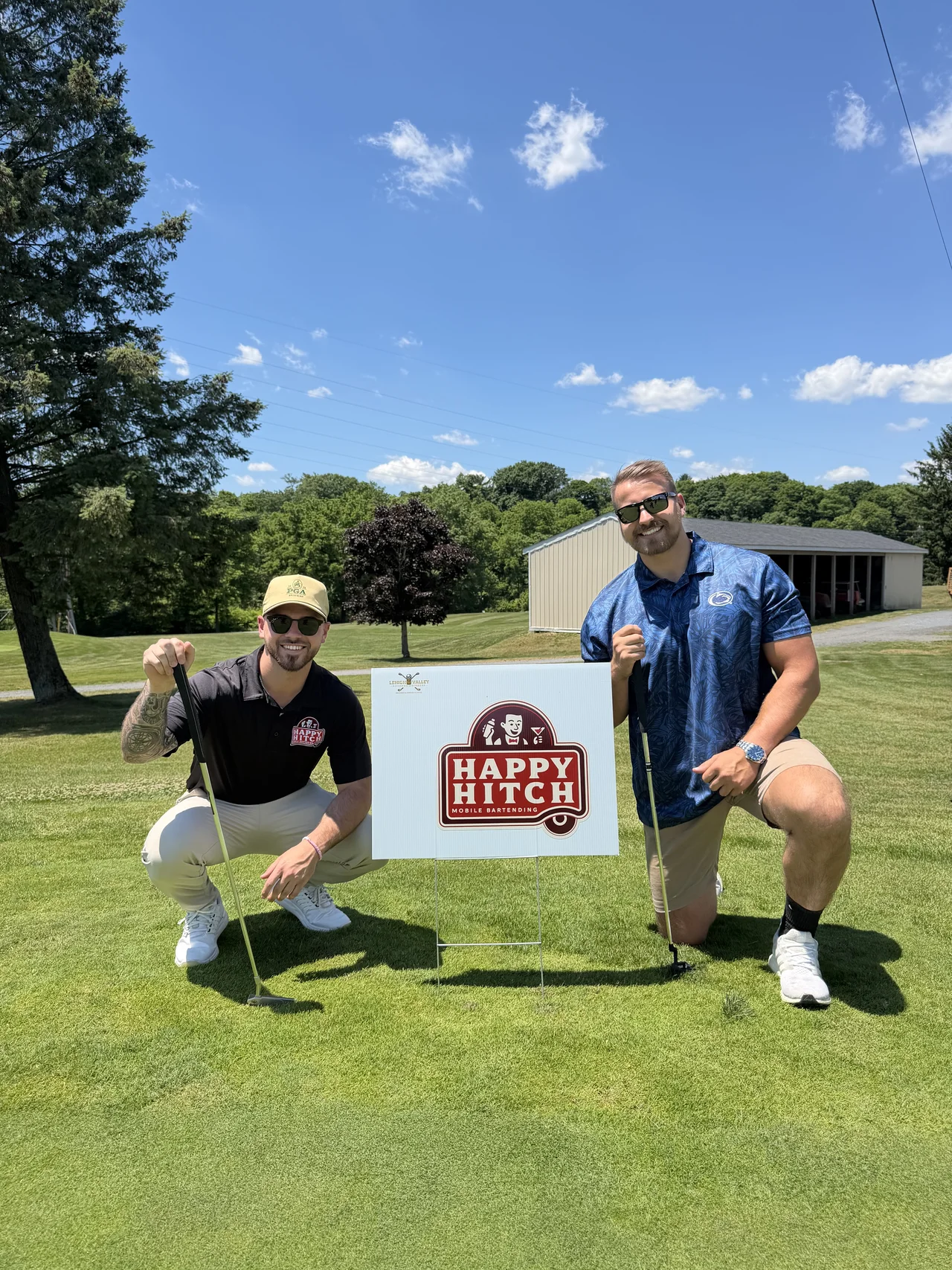Two people beside a Happy Hitch sign on a sunny lawn with trees in the background