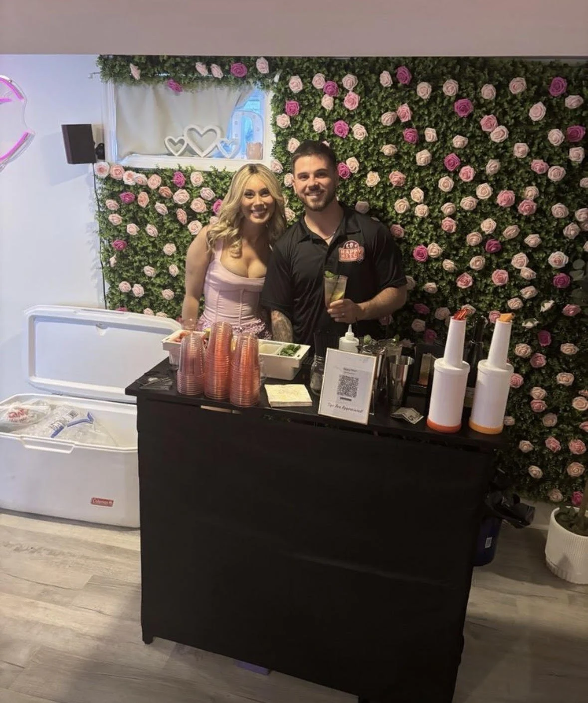 A bartender and a guest smiling in front of a pink rose wall at a black pop-up bar with cups, a cooler and a bar menu