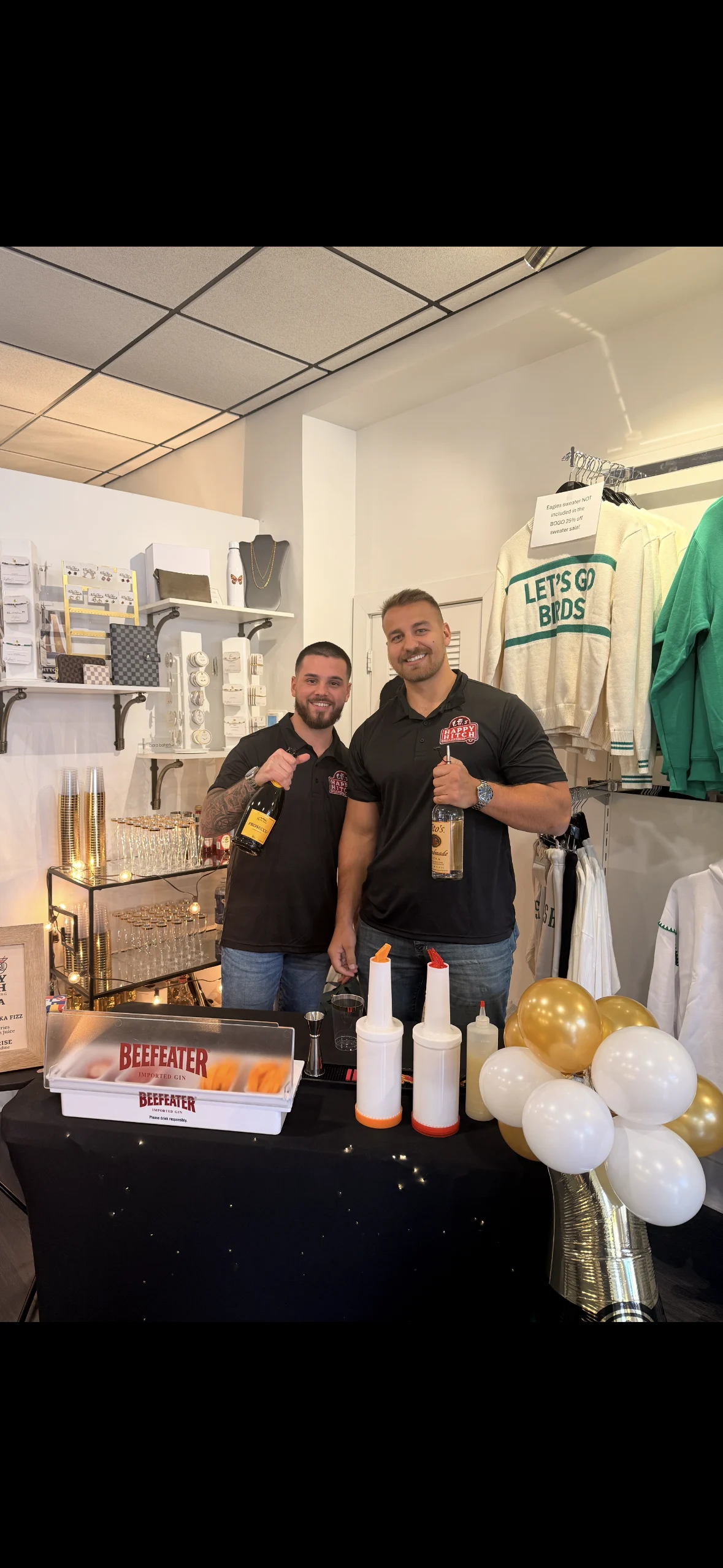 Jake and Dillon smiling behind a black pop-up bar with bottles, mixers and gold and white balloons in an indoor space