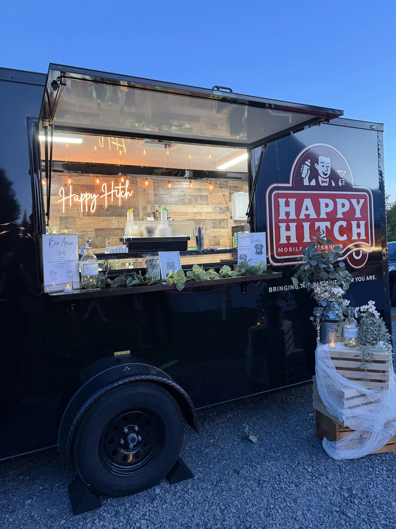 Black Happy Hitch bar trailer at dusk with its service window open, a neon sign, bar menus and flowers on a wooden crate beside it
