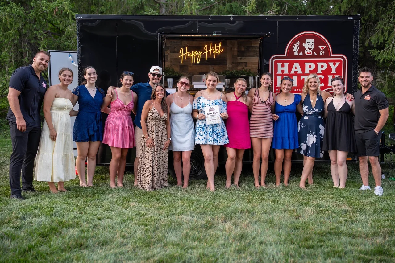 A group of guests and bartenders gathered in front of the open Happy Hitch trailer on a grassy lawn