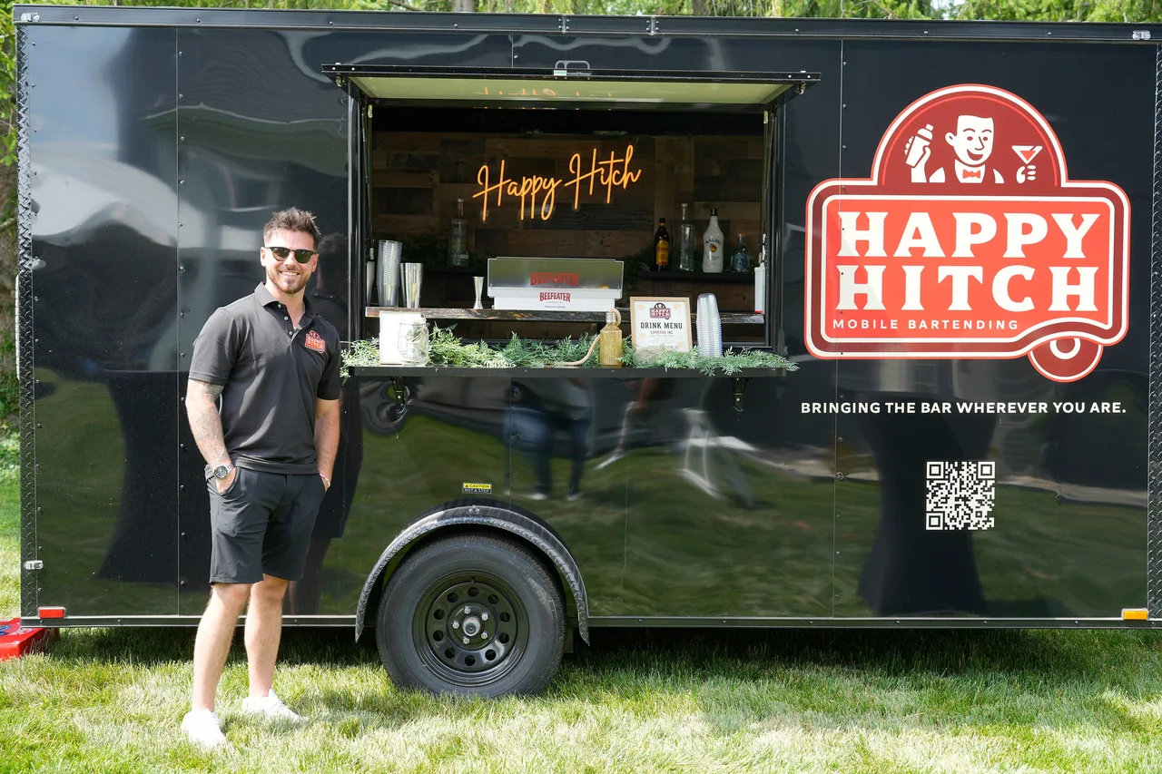 A Happy Hitch bartender beside the black trailer with its service window open and large red logo in daylight